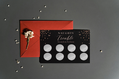 Hands holding a Naughty Trouble scratch card challenge with BDSM and role-play themes for adventurous couples.